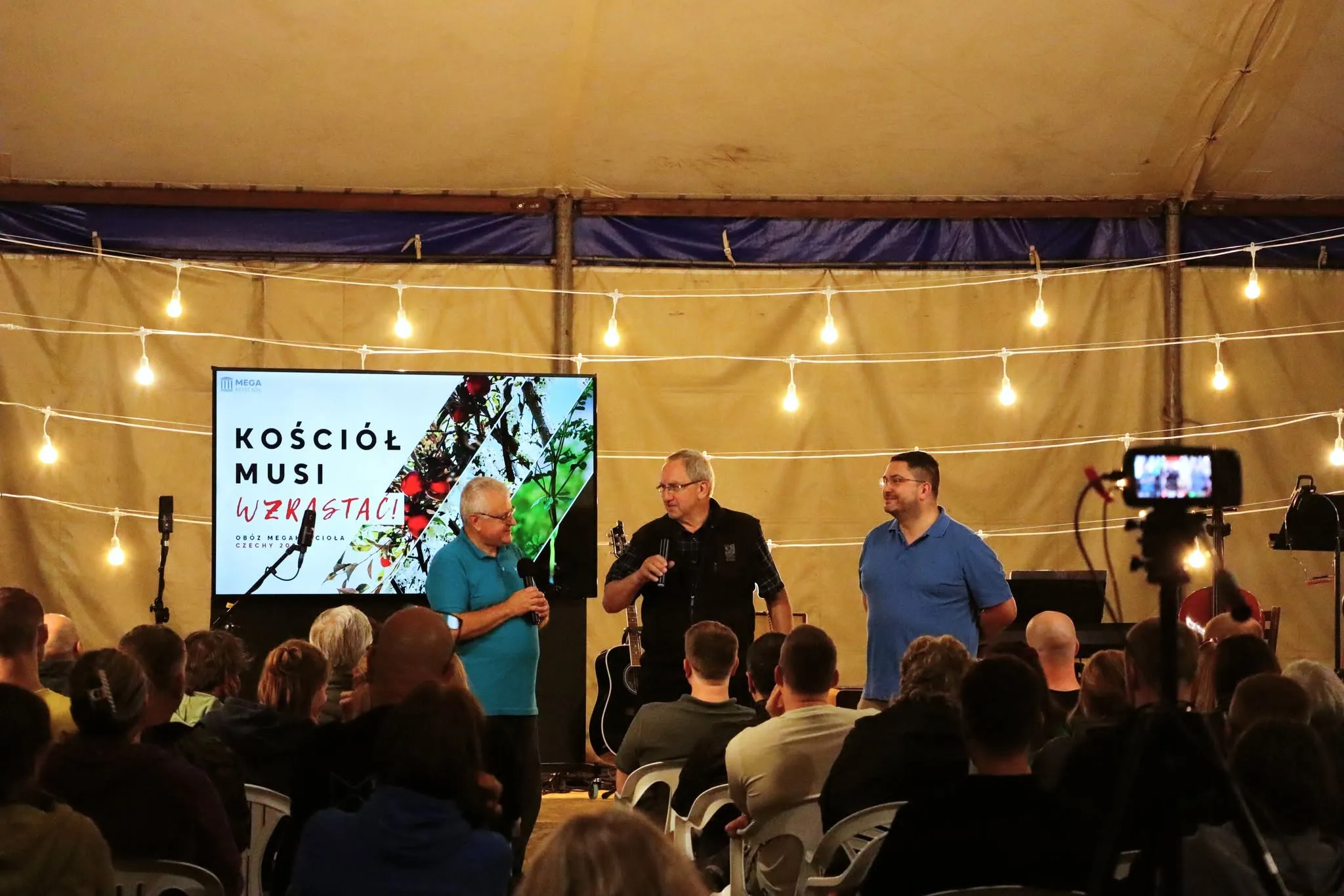 Trzech mężczyzn stoi na scenie w namiocie obozowym; na ekranie napis „Kościół musi wzrastać”.